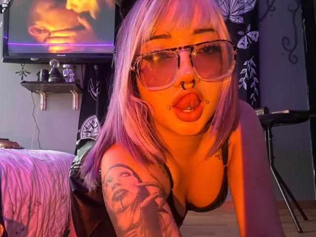 AgathaLin, avec ses cheveux violets, son anneau au nez, ses piercings aux lèvres et ses lunettes, portant un soutien-gorge noir, penchée en avant, montrant ses seins, et un tatouage sur son bras droit, dans une pièce éclairée en violet..