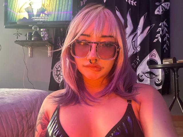 AgathaLin, avec ses cheveux violets et roses, portant de grandes lunettes de soleil, un haut noir brillant, un anneau au nez et un piercing aux lèvres, exhibant sa poitrine et son bras tatoué. Décor d'une chambre, éclairage coloré..