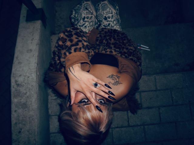 AgathaLin, con i capelli biondi, una giacca a stampa leopardo, unghie nere, un tatuaggio a forma di teschio e stivali con teschio, seduta sulle scale, con la mano a coprire il viso, illuminazione scura e malinconica..