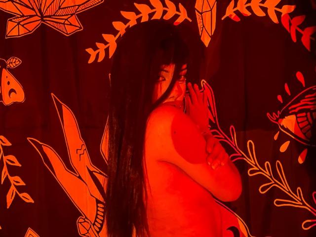AgathaLin met lang donker haar, ongetooid, waarbij ze haar borsten bedekt met haar hand, verlicht in rood-oranje licht, staand voor een donkere, patroonrijke achtergrond met blad- en vlinderontwerpen..