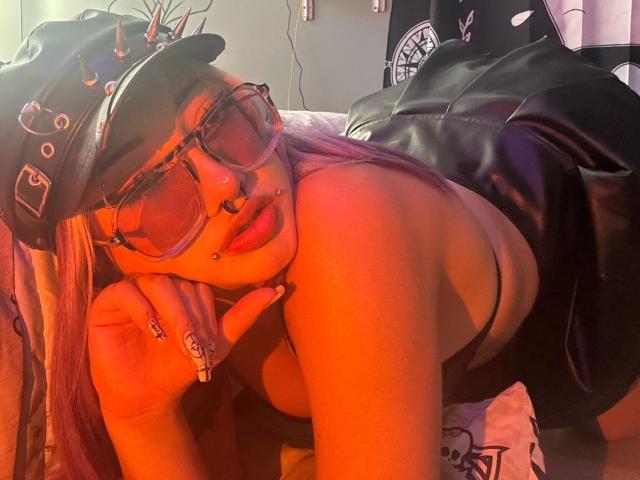 AgathaLin with pink hair, black cap, nose ring, and glasses, leans on her hand, showing her cleavage in a black top, with a black leather jacket behind her.