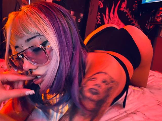AgathaLin, com cabelo roxo e branco, óculos, piercing no nariz e tatuagens, ajoelhada, vestindo uma calcinha preta, mostrando o bumbum, mordendo o dedo, em um quarto iluminado por luz vermelha..
