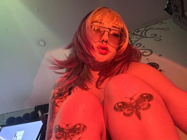 AgathaLin with pink and blonde hair, glasses, nose ring, and red lighting, showing tattooed butterflies on her knees, her big tits in front, and pouty red lips.