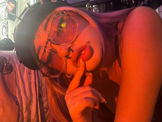 AgathaLin, avec ses cheveux roses, ses lunettes et son anneau de nez, suce son doigt la langue dehors, portant une casquette noire, baignée d'une lumière rouge-orange. Ses seins sont visibles..