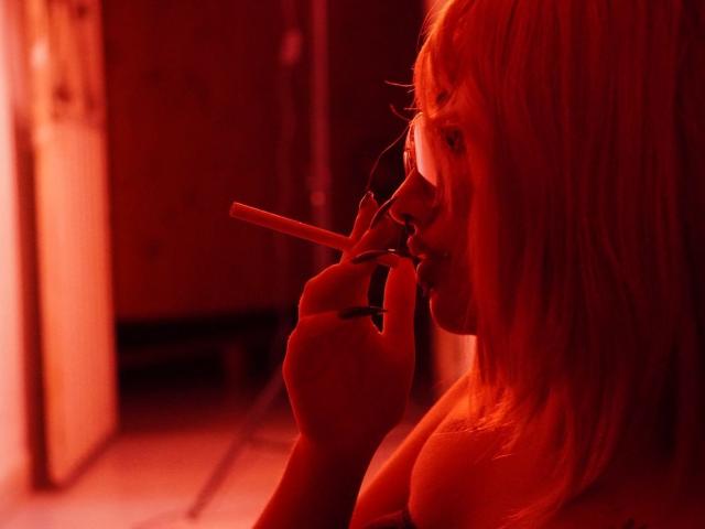 AgathaLin, con la piel iluminada en rojo, cabello rubio y senos pequeños, sosteniendo un cigarrillo, mordiendo la punta, mirando hacia un lado, en un entorno oscuro, melancólico y con tonos rojos..
