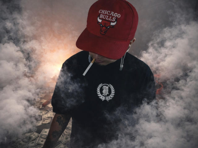 ZacWatson with a cigarette in his mouth, wearing a red Chicago Bulls cap and black shirt, surrounded by thick white smoke, with money on the ground.