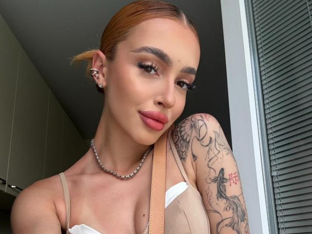MarissaFex with orange hair, makeup, and tattoos, wearing a white top and pearl necklace, showing her tits, with a confident smile, in a selfie.