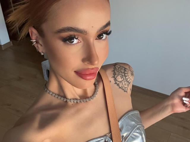 MarissaFex with blonde hair, brown eyes, and pink lips, wearing a silver top, necklace, and showing a tattoo on her right shoulder, holding a strap.