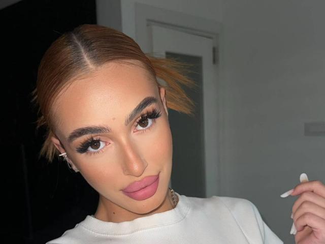 MarissaFex has light brown hair, full eyebrows, long eyelashes, pink lipstick, white shirt, and is holding a white object near her mouth. She's looking at the camera.
