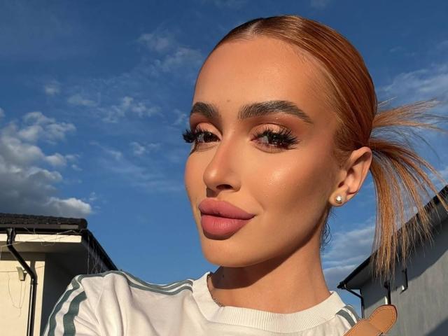MarissaFex with orange ponytail, wearing white Adidas, has glossy pink lips, long eyelashes, and diamond earrings, smiling under bright blue sky with fluffy clouds.