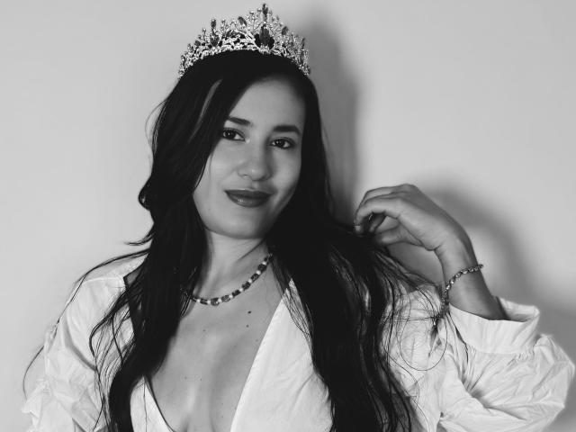 SarahMoonOne, com cabelos longos e pretos, usando uma coroa adornada, camisa branca, decote profundo, colar de contas e pulseira, sorrindo levemente, tocando o cabelo. Foto em preto e branco..