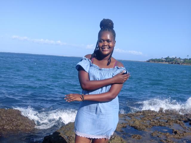 SeleneDarlyne with dark skin, medium-sized tits, and a cute smile, wearing a blue off-shoulder dress, standing on rocky shore, arms crossed, blue ocean in background.