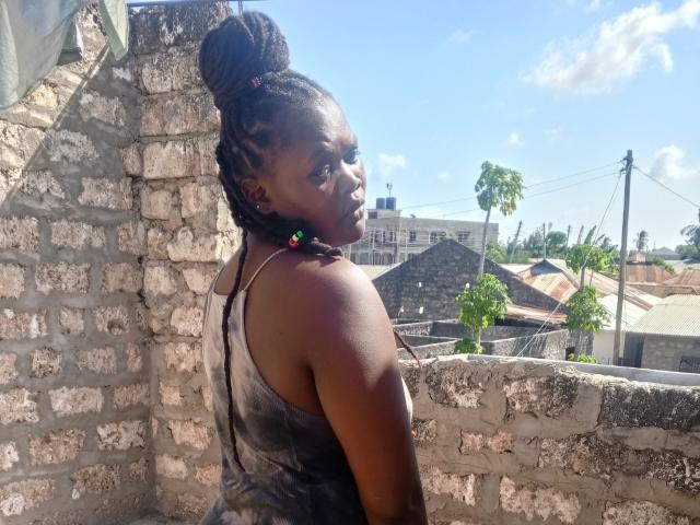 SeleneDarlyne, de piel oscura, con un moño trenzado, vestida con una blusa negra de estampado tie-dye, de pie frente a una pared de piedra, mirando por encima del hombro, con la luz del sol en la espalda, y pequeños aretes de colores..