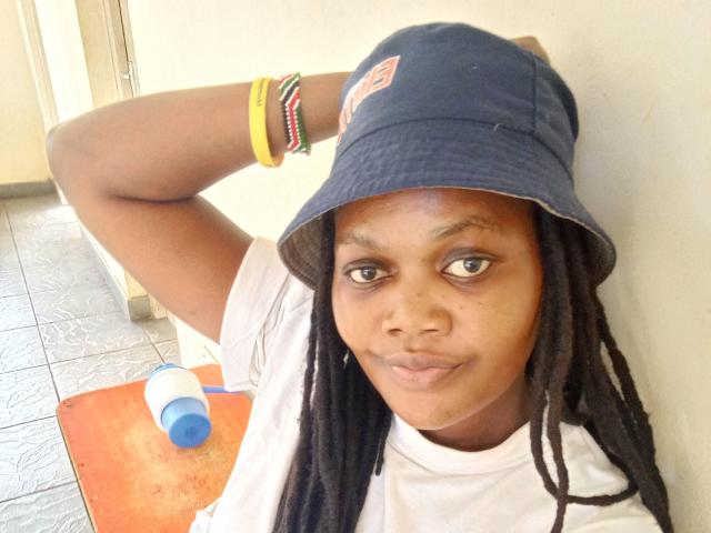 SeleneDarlyne, com pele escura, olhos castanhos e dreadlocks pretos, usando um boné azul, uma camisa branca e pulseiras coloridas, franzindo os lábios, segurando uma garrafa azul e branca..