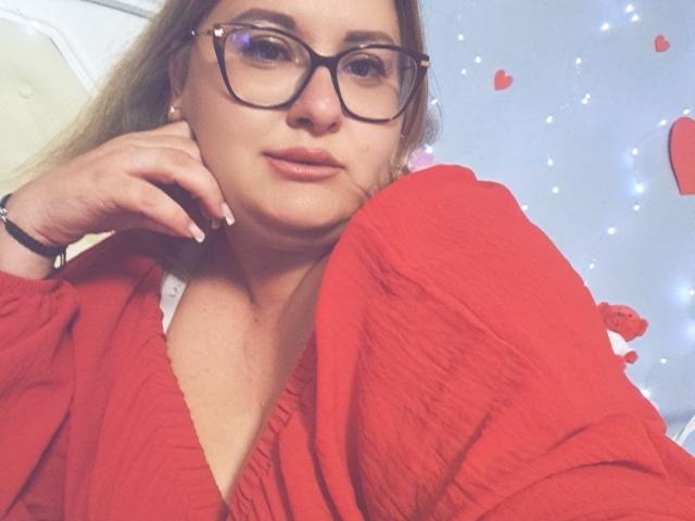 RadaRori with glasses, red ribbed shirt, and a slightly pouty expression, resting her chin on her hand, showing off her subtle makeup and long blonde hair.