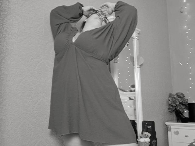 RadaRori porte une robe ample à manches longues avec une fente sur le devant, les bras levés, la tête penchée en arrière, laissant entrevoir un peu de ses seins et un bracelet en pompon duveteux. Photo en noir et blanc..
