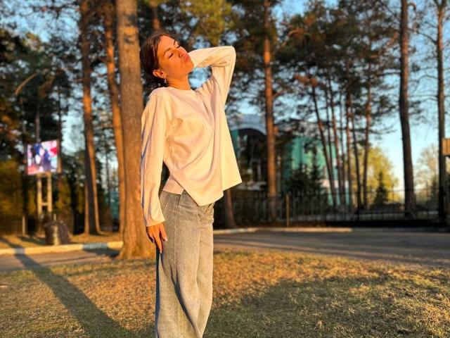 RoxanaKrausse with eyes closed, hand in hair, wearing loose white shirt and gray pants, standing in sunlit park, holding blue water bottle, relaxed, tall trees in background.