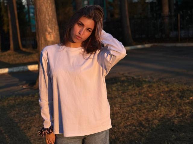 RoxanaKrausse stands in a park, wearing a loose white shirt and blue jeans, with her hand in her brown hair, sunlight casting a warm glow on her.
