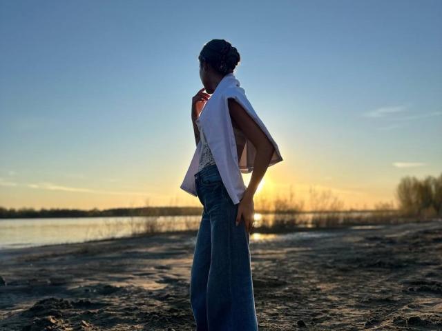 RoxanaKrausse stands on a sandy shore, backlit by sunset. She's wearing a white sleeveless top, blue jeans, and has a small, round butt. Her hair is in a bun.