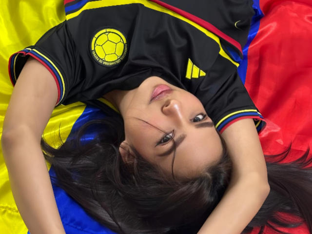 Sensuelle Camila mit dunklem Haar, liegt auf dem Rücken, trägt ein schwarzes Fußballtrikot mit gelben und roten Akzenten, die Arme sind über dem Kopf, sie schaut in die Kamera, die Augen sind halb geöffnet..