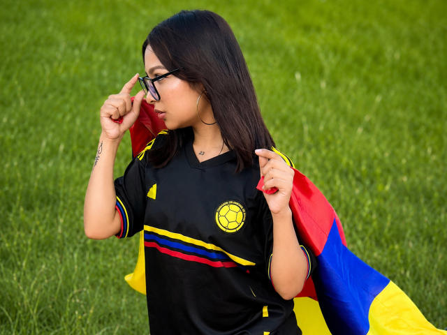 SensualCamila with black soccer shirt, yellow and red stripes, and soccer logo, wearing glasses, adjusting them, standing with Bolivian flag over her shoulder, green grass background.