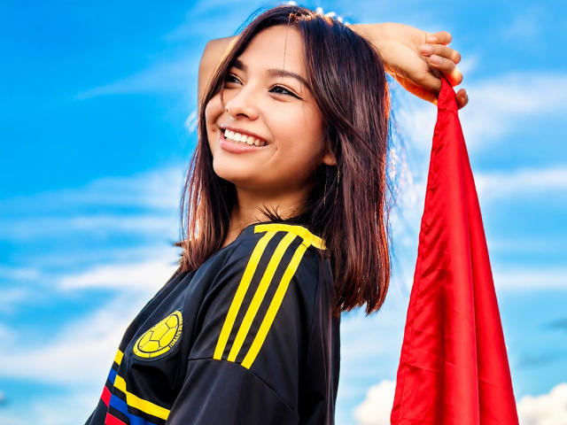 Sensual, Camila, com cabelo castanho, sorrindo, segurando uma bandeira vermelha acima da cabeça, vestindo uma camisa de futebol preta com listras amarelas e um logotipo amarelo, contra um céu azul brilhante..