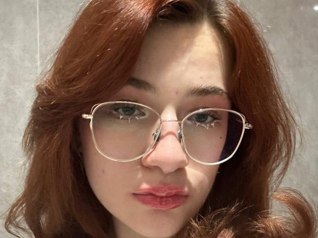 Isabel Barfuss, con i capelli rossi, indossa occhiali trasparenti, ha gli occhi azzurri e le labbra rosa. Il suo naso è piccolo e guarda direttamente la fotocamera con un'espressione neutra..