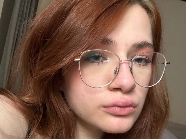 Isabel Barfuss, met rood haar, die ronde brillen draagt, heeft een kleine tatoeage in de buurt van haar rechteroog en volle, roze lippen. Haar groene ogen en lichte huid vallen op..