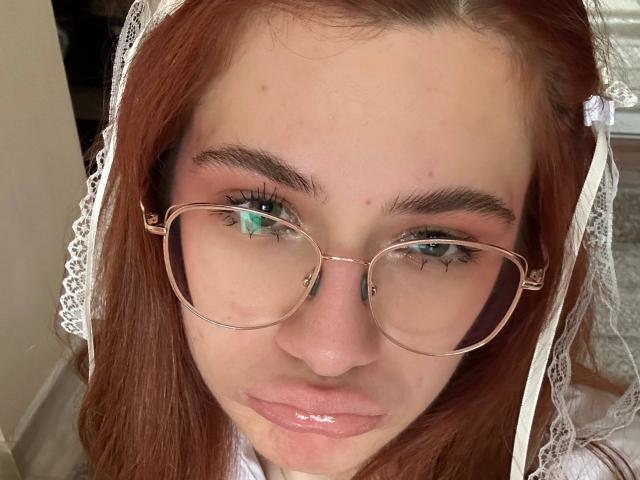 Isabel Barfuss, met glanzende roze lippen, draagt ronde brillen en een witte kanten sluier, heeft rood haar en groene ogen, en kijkt recht in de camera met een lichte frons..