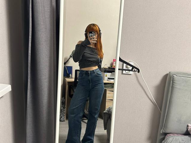 Isabel Barfuss, met rood haar, draagt een koptelefoon, een gestreept topje en hoge jeans, en maakt een spiegelzelfportret waarop haar achterwerk en jeans te zien zijn. Reflectie in een slaapkamer..