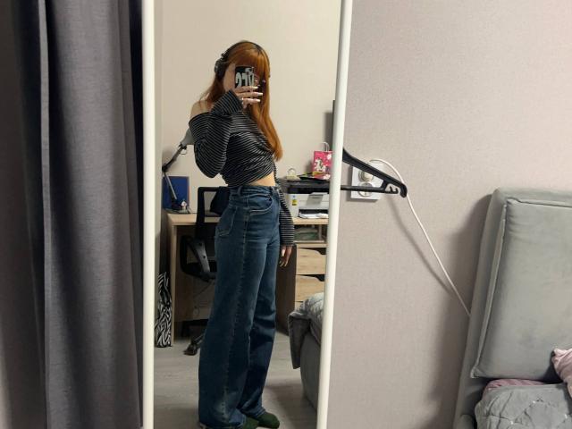 Isabel Barfuss, met lang rood haar, draagt een zwart-wit gestreept topje en een blauwe spijkerbroek met hoge taille, en staat voor een spiegel, waarbij haar billen en borsten zichtbaar zijn..