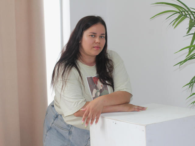 LoraKrei, com cabelos longos e pretos, vestindo uma camiseta branca e calças jeans azuis de cintura alta, apoiada em um cubo branco, com uma expressão levemente irritada, e uma planta verde à direita..