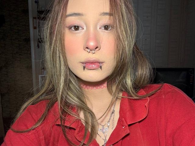 Siobhan Sunderland tiene un piercing en la nariz, piercings en los labios y un collar rosa. Lleva una camisa roja y un collar con cuentas moradas. Su cabello castaño está suelto..
