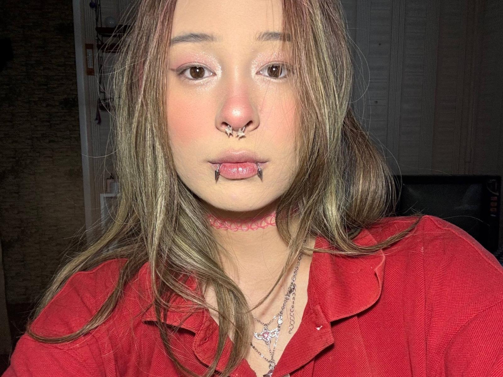 Siobhan Sunderland tiene un piercing en la nariz, piercings en los labios y un collar rosa. Lleva una camisa roja y un collar con cuentas moradas. Su cabello castaño está suelto..