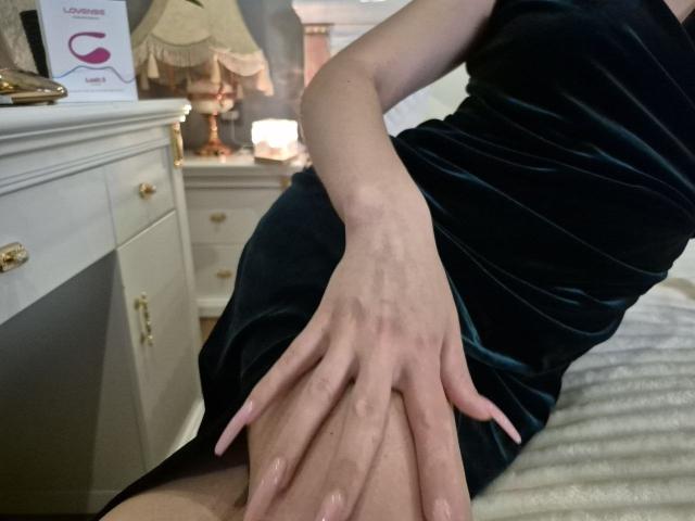 Rouzi sitzt in einem schwarzen Samtkleid, ihre Hände ruhen auf ihrem Hintern, wobei blasse Haut und lange, rosafarbene Nägel zu sehen sind, in einem sanft beleuchteten Schlafzimmer..