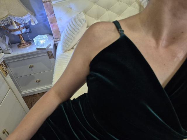 Rouzis schlanker Arm in einem schwarzen Samtkleid mit grünen Streifen, das ihre Schulter und ihren Schlüsselbein zeigt, neben einem weißen Bett mit einem gepolsterten Kopfteil..