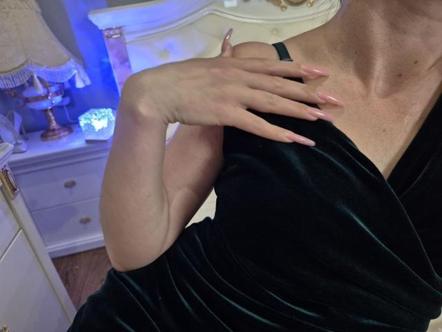 Rouzis Hand mit den langen, rosafarbenen Nägeln berührt ihr schwarzes Samtkleid und zeigt ihren Schlüsselbein und ihre glatte Haut. Ihre Brüste sind leicht unter dem Kleid sichtbar..