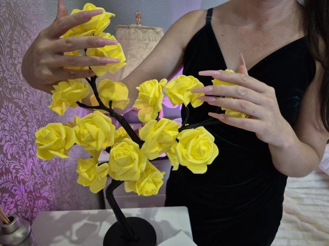 Rouzi trägt ein schwarzes Kleid und hält einen Strauß gelber Rosen mit beiden Händen. Ihre Nägel sind rosa lackiert, die Blumen haben einen schwarzen Stiel, und das ganze befindet sich in einem sanft beleuchteten Raum..