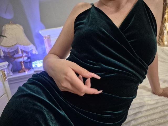 Rouzi trägt ein dunkelgrünes Samtkleid, ihre Hand ruht auf ihrer weichen, runden Brust, sie sitzt auf einem weißen Bett, und ihr langes, braunes Haar ist auf der rechten Seite sichtbar..