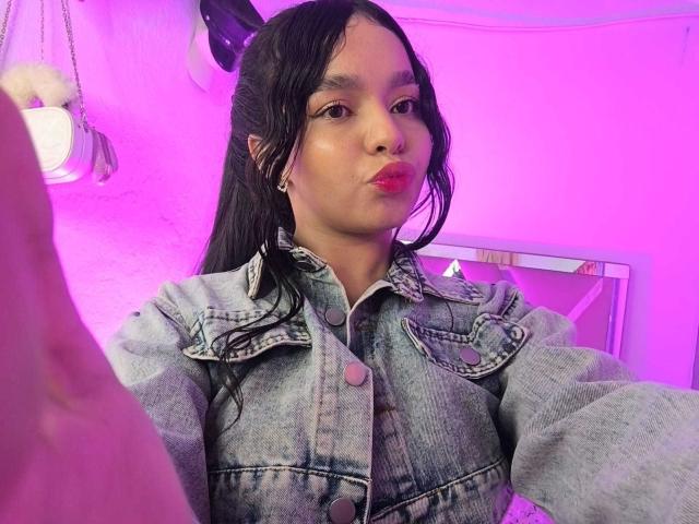 Lissy Carter, con cabello oscuro recogido en una coleta, vistiendo una chaqueta vaquera, con los labios rosados fruncidos, mirando a la cámara, habitación iluminada con luz púrpura, pequeños aros, pintalabios rosa visible..