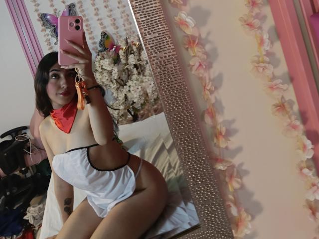 AngelOh, avec des cheveux bruns, une écharpe rouge, de la lingerie blanche sans bretelles, tenant un téléphone rose avec des autocollants de chats, un selfie devant un miroir, montrant ses fesses nues, des fleurs roses et un tatouage sur sa cuisse gauche..