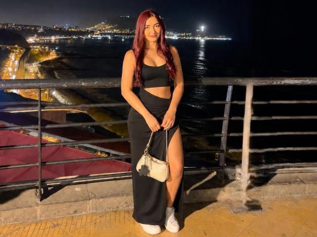 SalomeJoe stands on a balcony at night, wearing a black crop top and skirt with high leg slit, holding a white purse, and has red hair, medium tits.