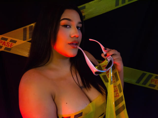 ValeryRiven, con i capelli scuri, illuminazione rossa, che tiene un dildo di plastica trasparente vicino alla bocca leggermente aperta, indossa un top giallo senza spalline, che rivela il suo seno generoso contro un nastro giallo di avvertimento..