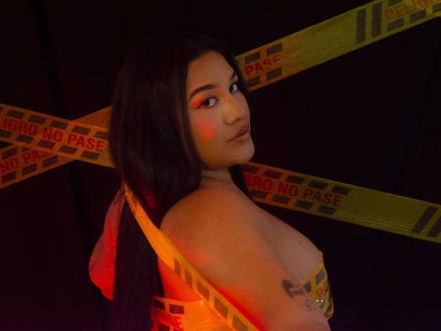 ValeryRiven mit langem, schwarzem Haar, die über die Schulter schaut, mit orange-roter Augenmakeup, gelbtem Band mit der Aufschrift "KEIN EINTRITT" über der Brust, nackten Schultern, Tattoo am rechten Arm, weiches Licht, dunkler Hintergrund..