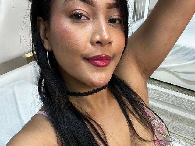 VickyRuiz, met donker haar, roze lippenstift, een zwarte choker, oorringen en een lichte glimlach, heft haar arm, toont haar borsten en heeft een subtiele gebruinde huid..