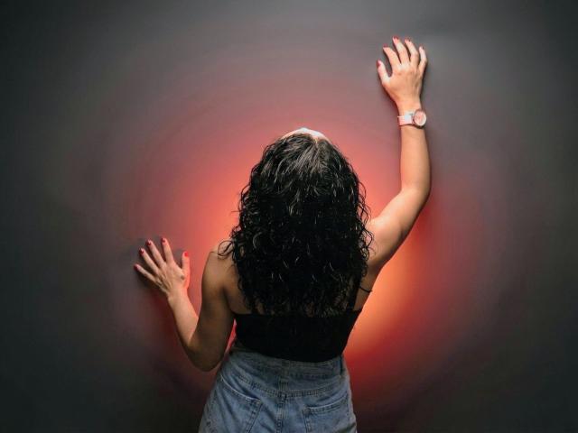 AlejaPierce's back is to the camera, hands against a dark wall, wearing a black top and blue jeans, with curly black hair and a pink wristwatch.
