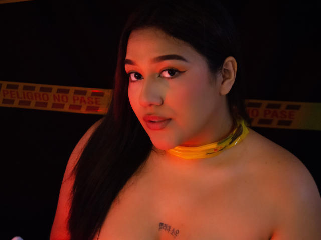 ValeryRiven with long black hair, yellow collar, and subtle makeup, topless with small tits, tattoo under right breast, looking at camera with red and orange lighting.