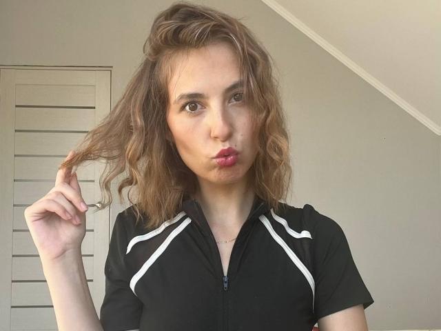 Tora Stambaugh, con cabello castaño ondulado, labios rosados y finos, vestida con una camisa negra con cremallera y rayas blancas, sosteniendo su cabello, mirando a la cámara..