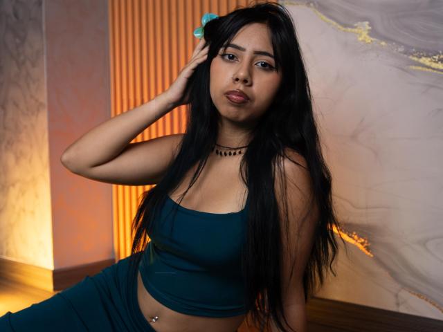 AyameFoxy with long black hair, dark green crop top, and matching pants, sitting, hand in hair, subtle belly button piercing, looking at camera, choker necklace, medium tits.