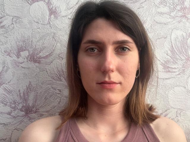 WanetaMalpica has pale skin, green eyes, brown shoulder-length hair, small black ear gauges, and wears a pink ribbed tank top. Her expression is neutral and she looks straight at the camera.
